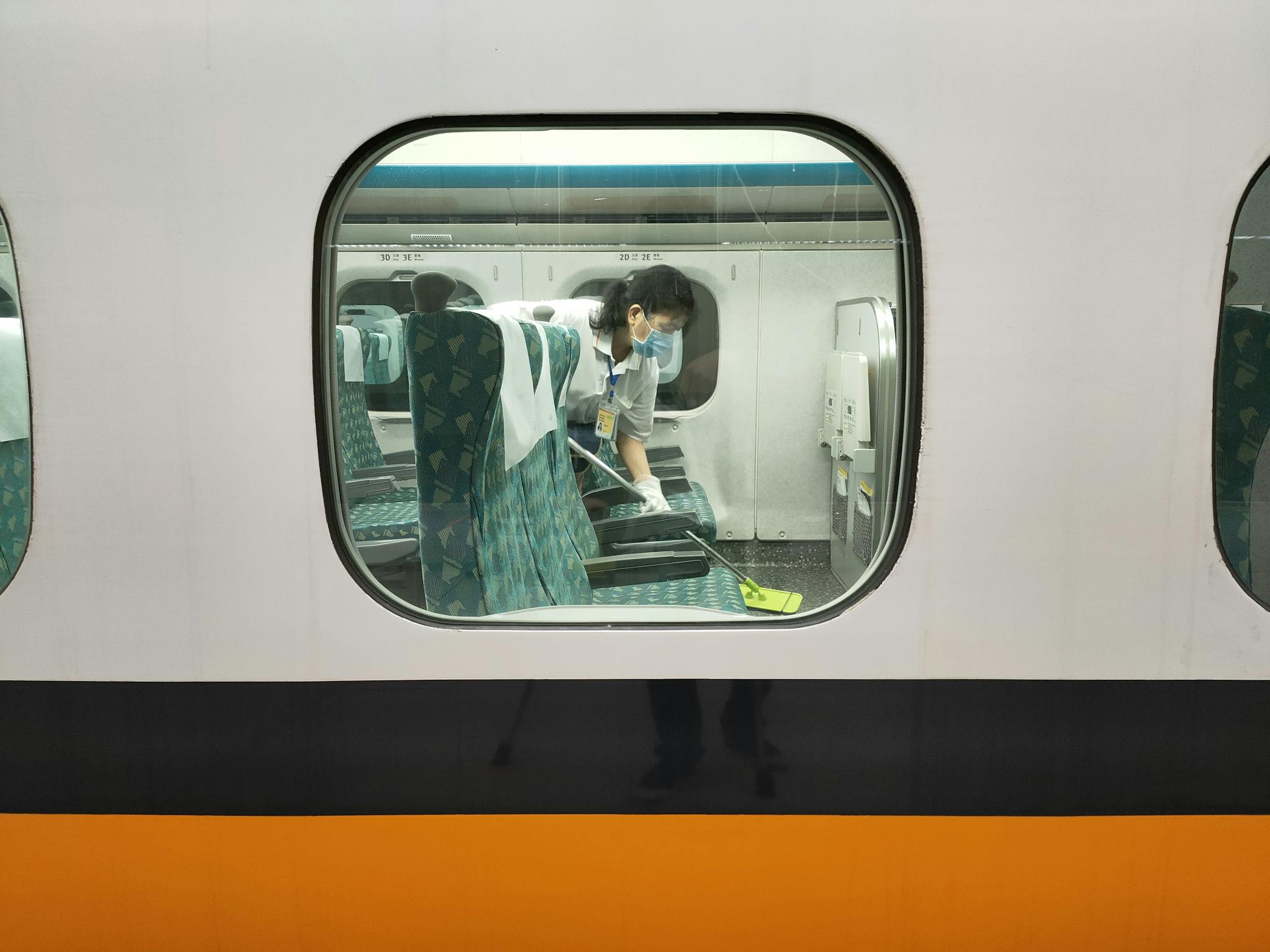 A woman in a face mask cleaning train seats with a mop, viewed through a window.