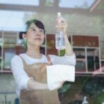 Asian woman wiping glass with a spray bottle and cloth, inside a restaurant setting.